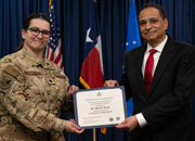 A person in military uniform and a man in a suit both face the camera holding a certificate