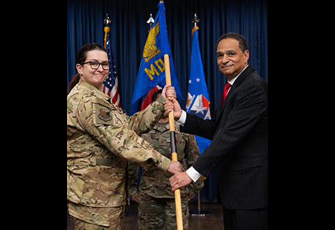 A person in military uniform and a person in a suit face the camera while holding a military guidon