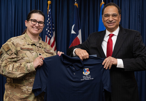 A person in military uniform and a person in a suit face the camera and hold a polo shirt
